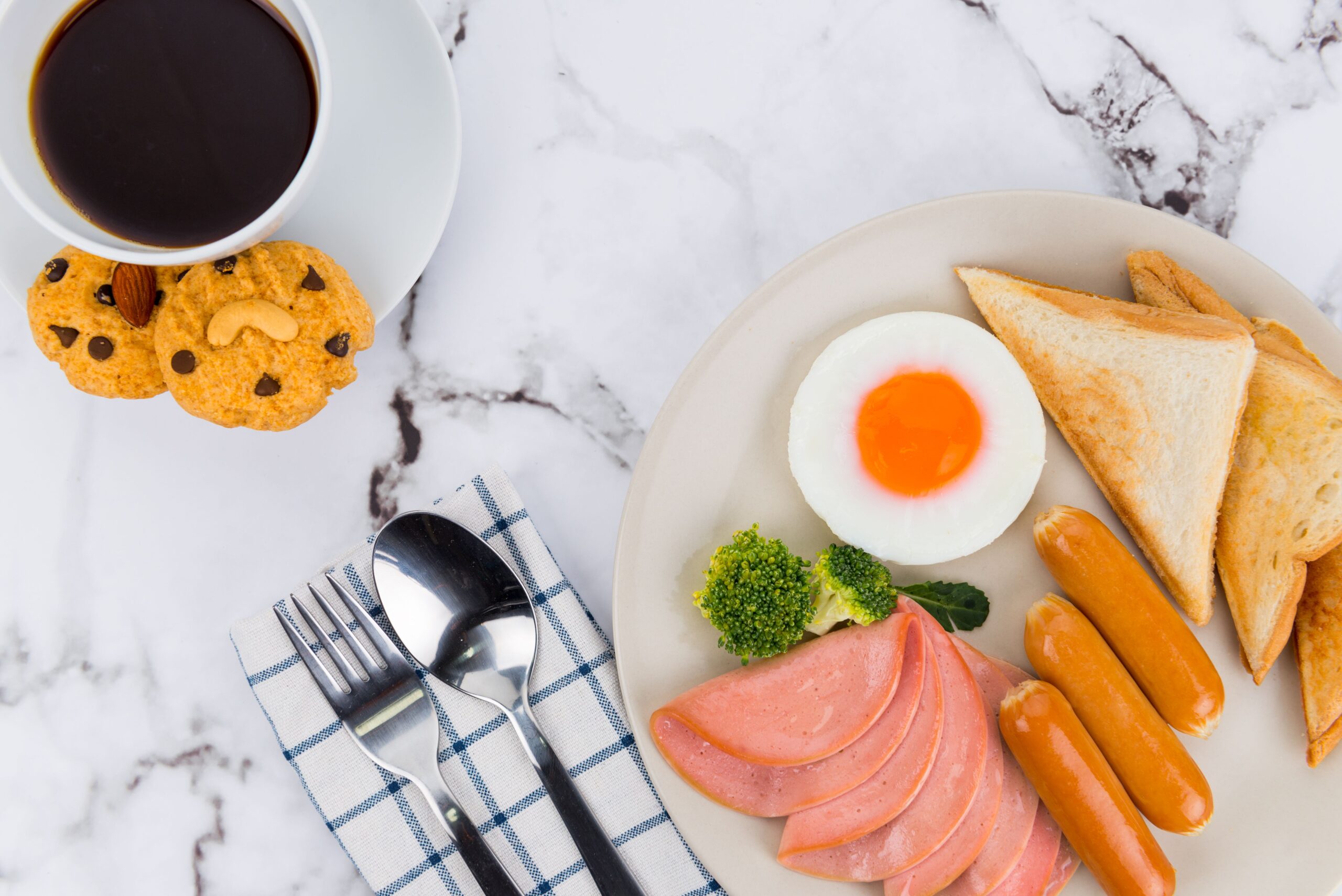 Healthy fitness breakfast with eggs, toast, sausage, sliced meat, broccoli, cookies, and black coffee on a marble table, representing balanced nutrition basics for fitness and muscle gain.